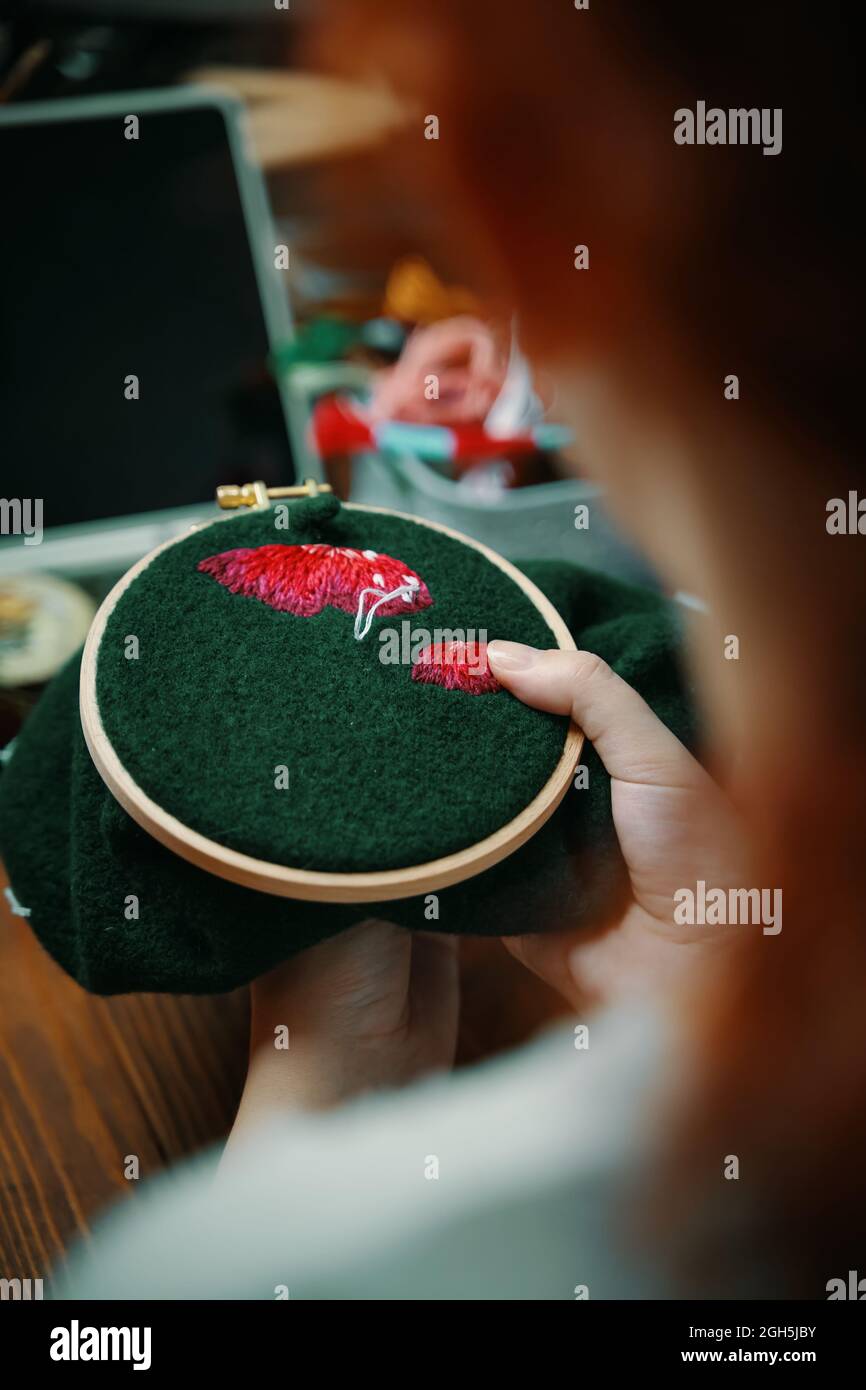 Cappello di fungo ricamato ragazza rossa in cerchio. Su materiale verde. Concetto di lavoro di ago, hobby, tempo libero. Primo piano. Tavolo in legno con computer portatile su sfondo. Foto Stock