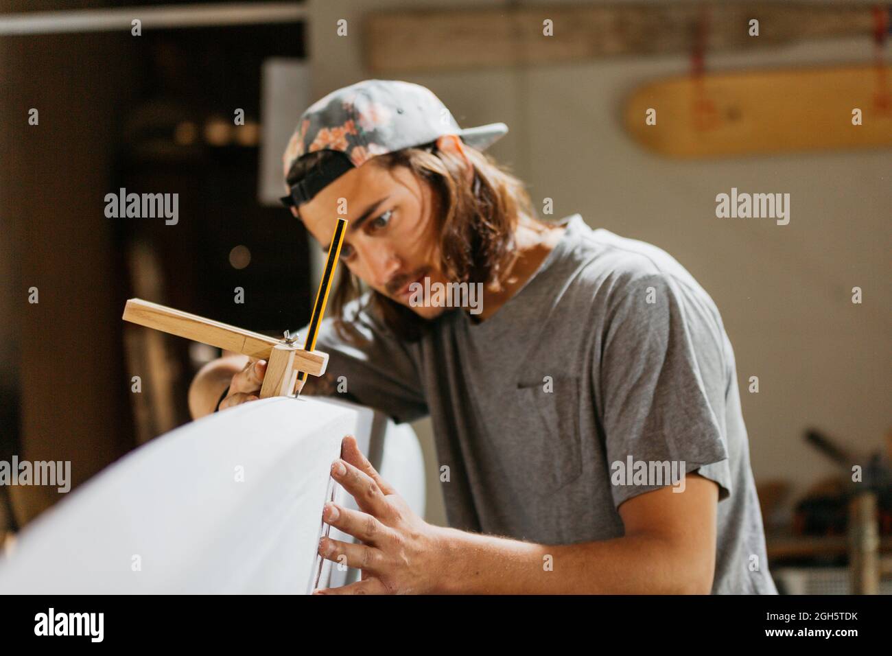 Hipster maschio concentrato usando l'attrezzo di incisione con la matita mentre marcano la tavola da surf prima di modellare in officina Foto Stock