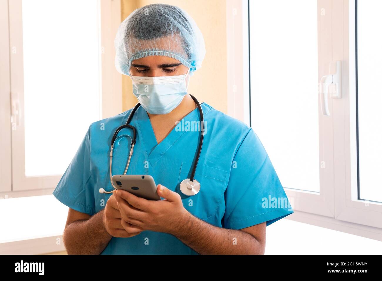 Medic maschile in uniforme e con stetoscopio in piedi in ospedale e navigare in Internet su smartphone Foto Stock