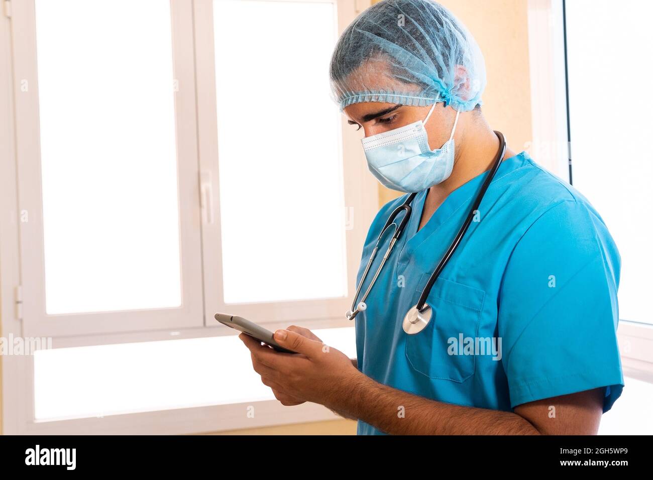 Medic maschile in uniforme e con stetoscopio in piedi in ospedale e navigare in Internet su smartphone Foto Stock