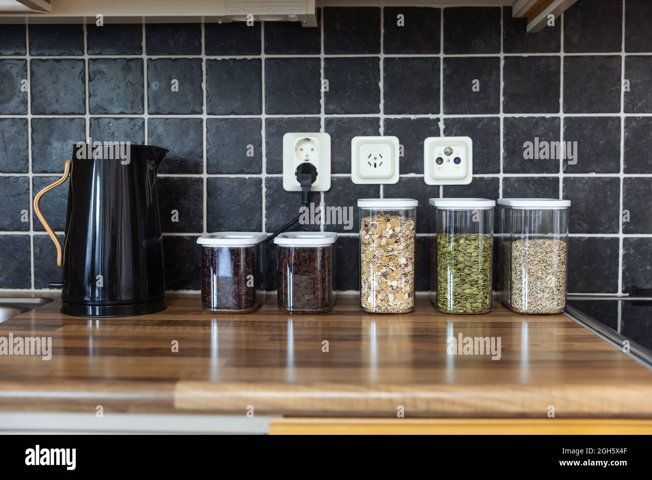 Contenitori di plastica con muesli e semi di girasole e zucca posti vicino ai chicchi di caffè e bollitore elettrico sul banco in cucina Foto Stock