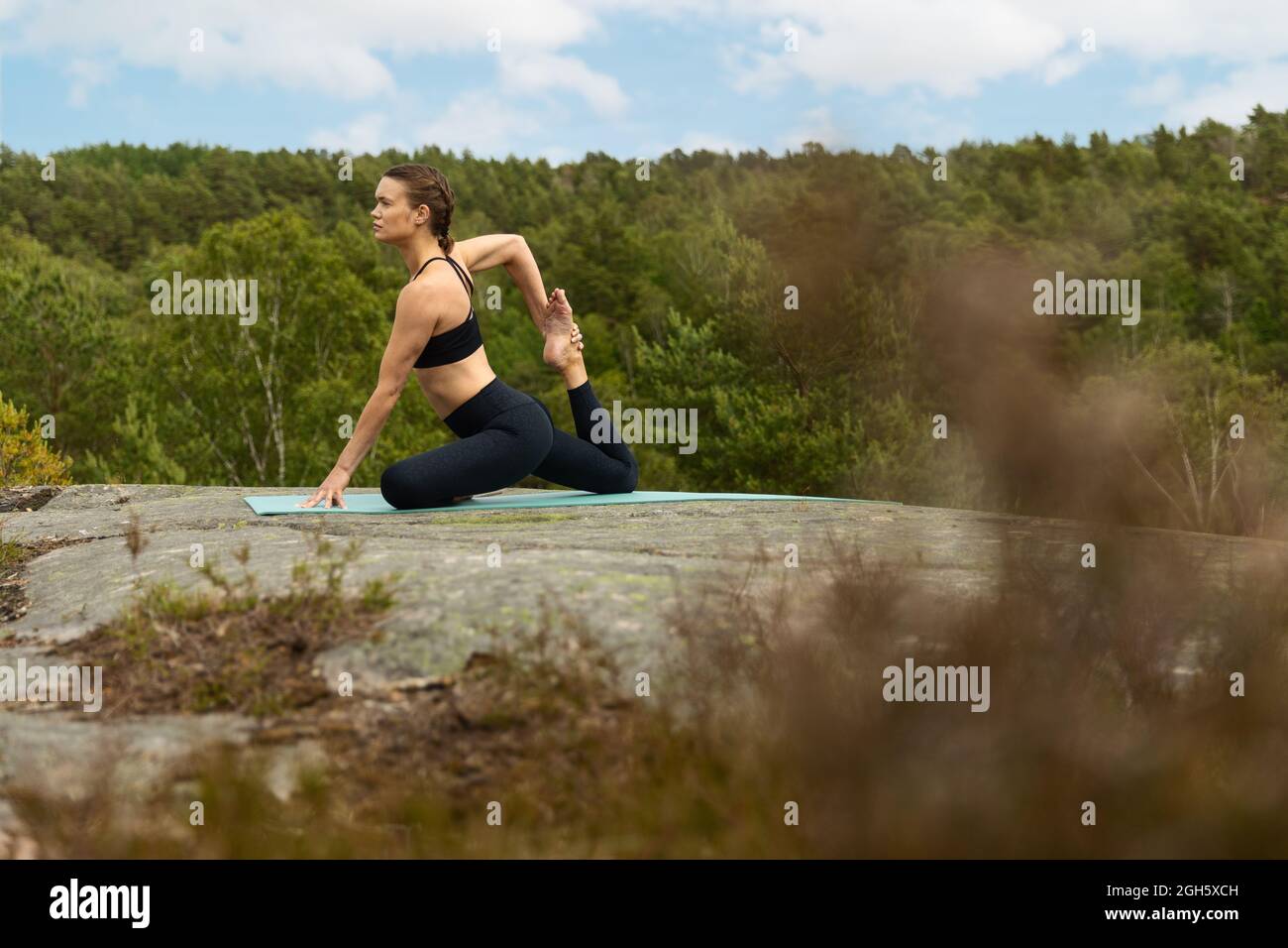 Sottile a piedi nudi femmina in nero activewear stretching corpo in Eka Pada Raja Kapotasana posa sulla pietra durante la sessione di yoga in campagna Foto Stock