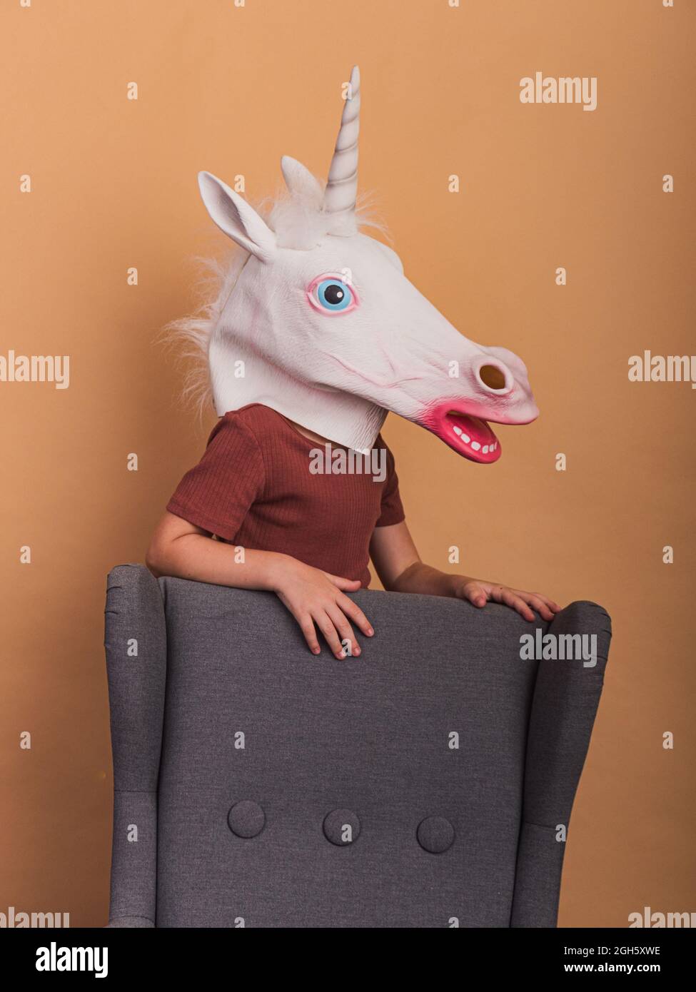 Capretto anonimo in maschera decorativa unicorno con bocca aperta poltroncina a contatto su sfondo beige Foto Stock