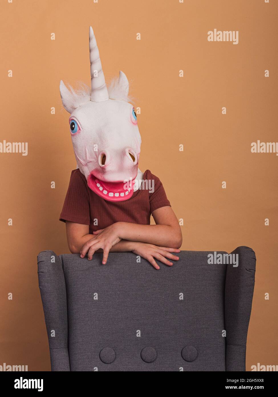 Capretto anonimo in maschera decorativa unicorno con bocca aperta poltroncina a contatto su sfondo beige Foto Stock