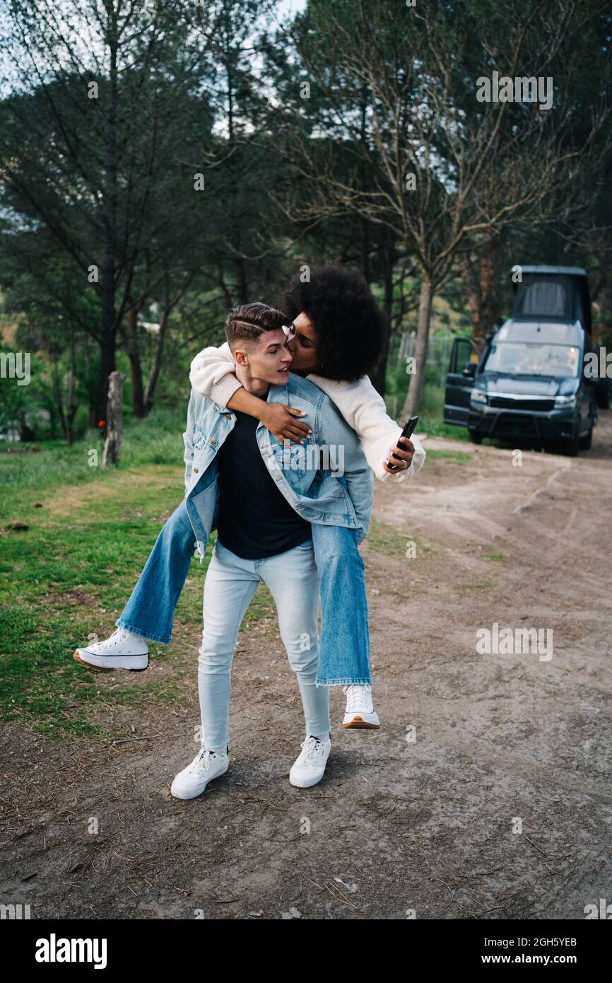 Giovane uomo che dà allegro etnia ragazza Piggyback giro contro camper e Thai Ridgeback sul sentiero Foto Stock