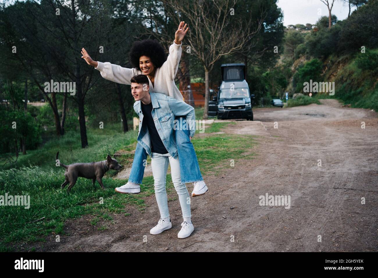 Giovane uomo che dà allegro giro di ragazza etnica piggyback mentre guarda via contro camper e Ridgeback tailandese sul sentiero Foto Stock