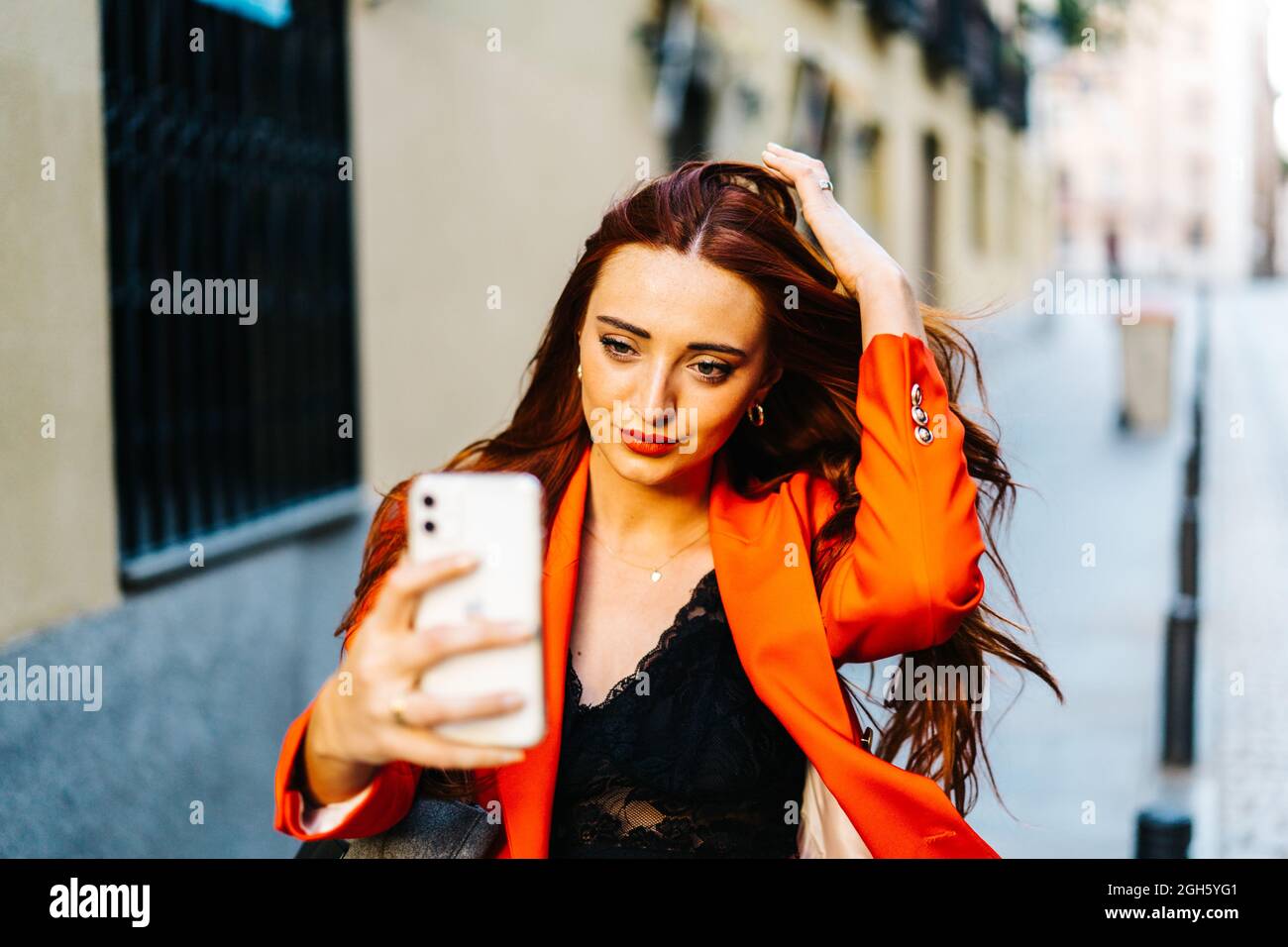 Donna elegante con capelli rossi e una vivace giacca arancione che scatta in auto sullo smartphone in città Foto Stock