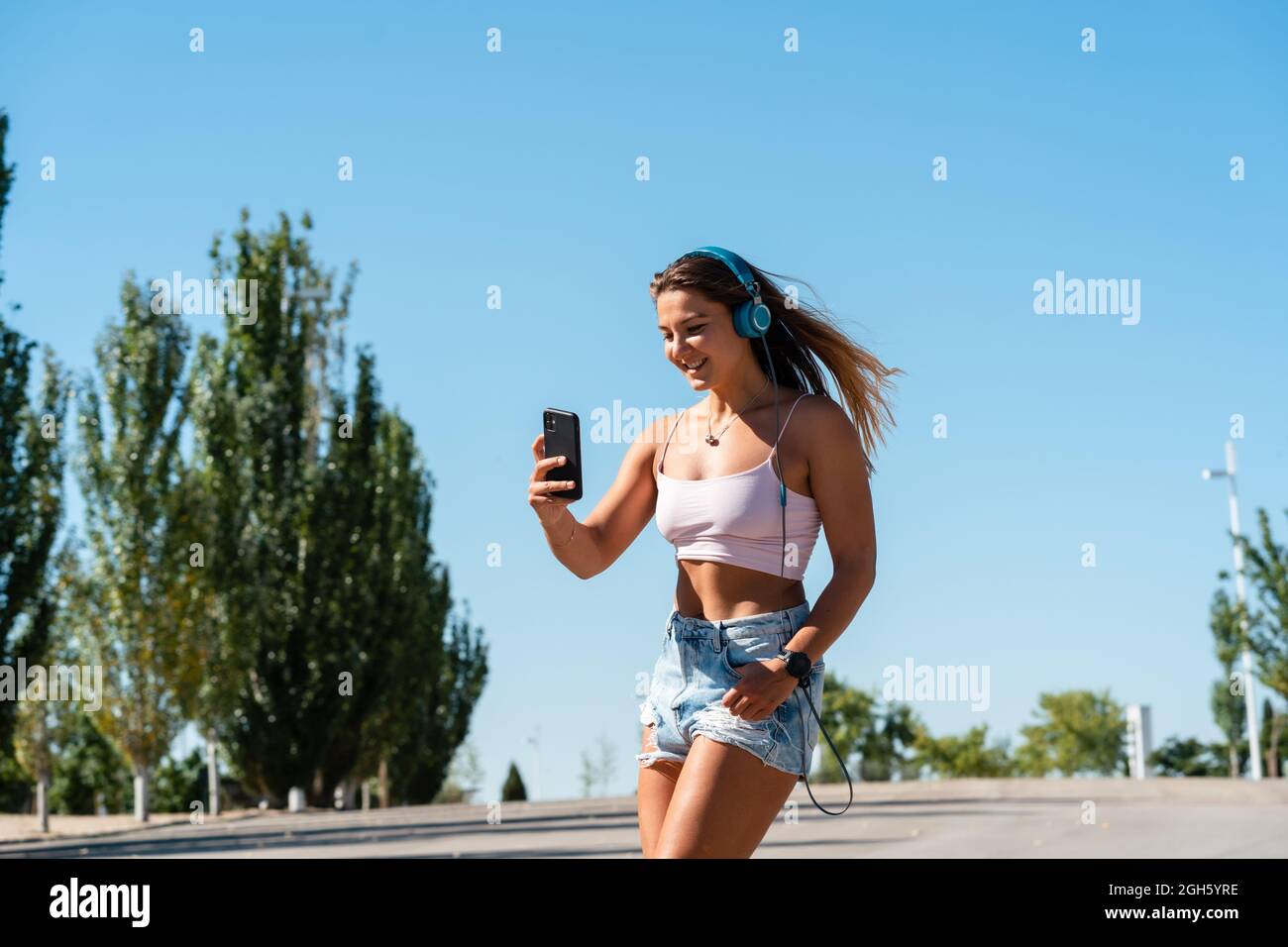 Positivo skater femminile in rollerblade e cuffie scattando auto-shot sul telefono cellulare il giorno di sole in estate in città Foto Stock