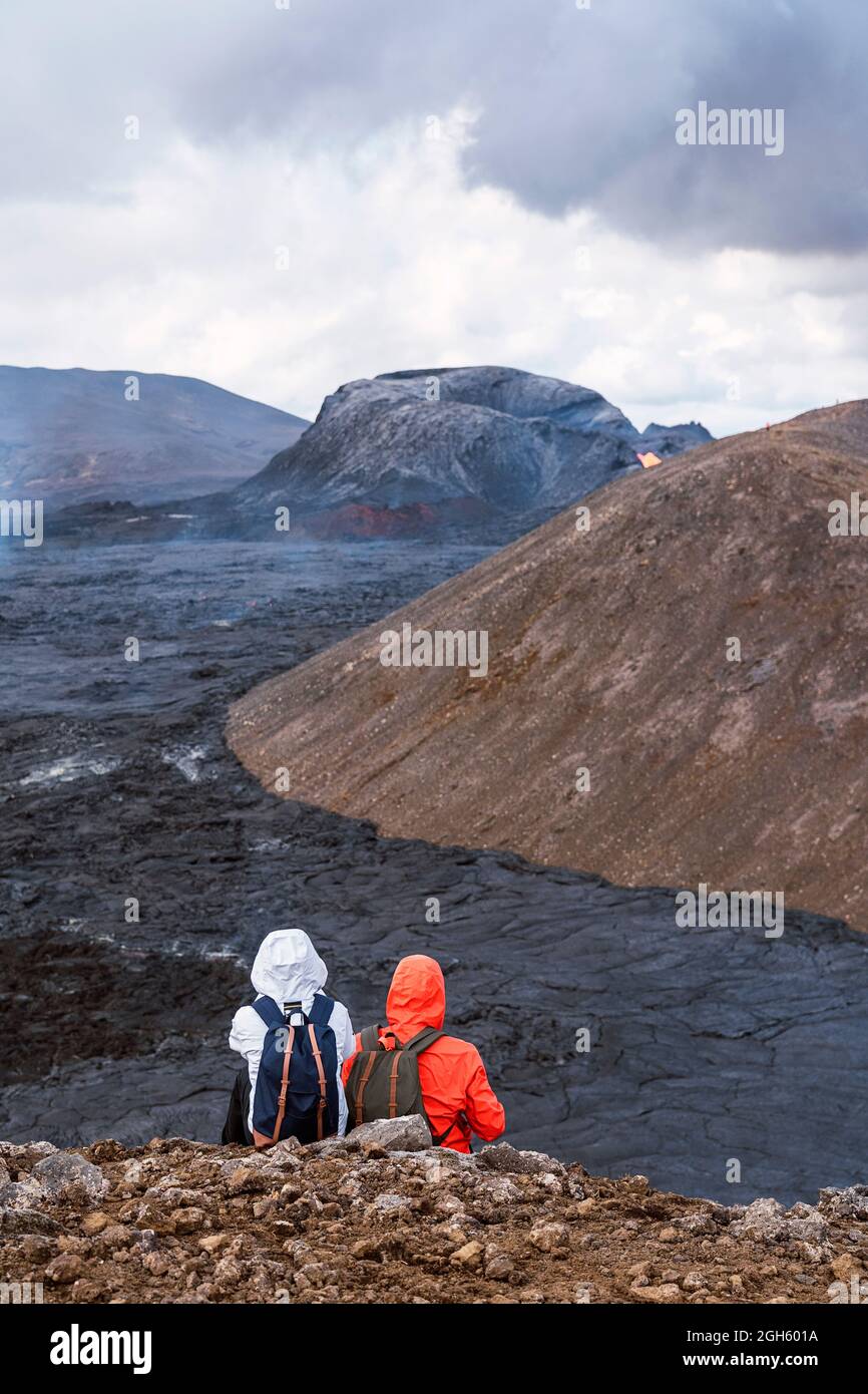 Vista posteriore di anonimi viaggiatori in outerwear contemplano la lava di Fagradersfjall dal monte sotto cielo nuvoloso in Islanda Foto Stock