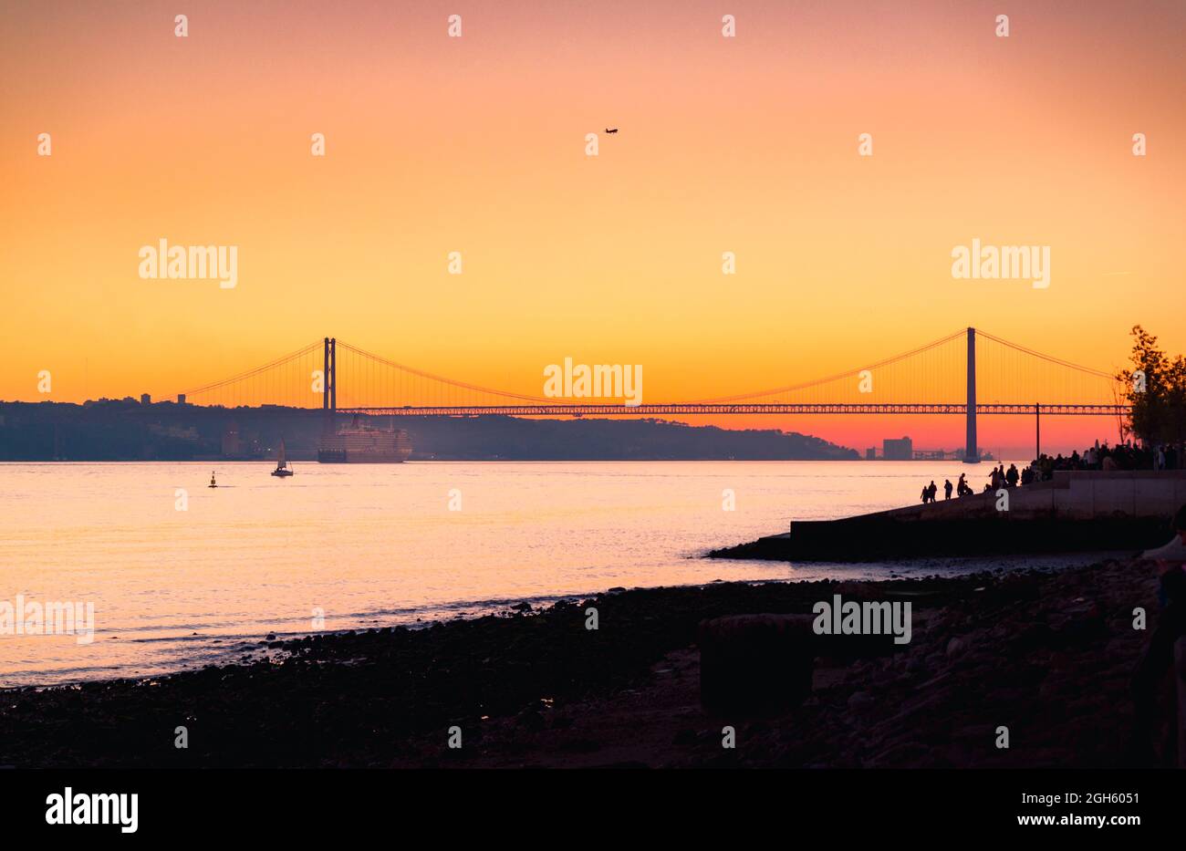 Argine affollate situate vicino al fiume Tago con traghetto vicino alla silhouette del ponte 25 de Abril contro il cielo arancione tramonto a Lisbona, Portogallo Foto Stock