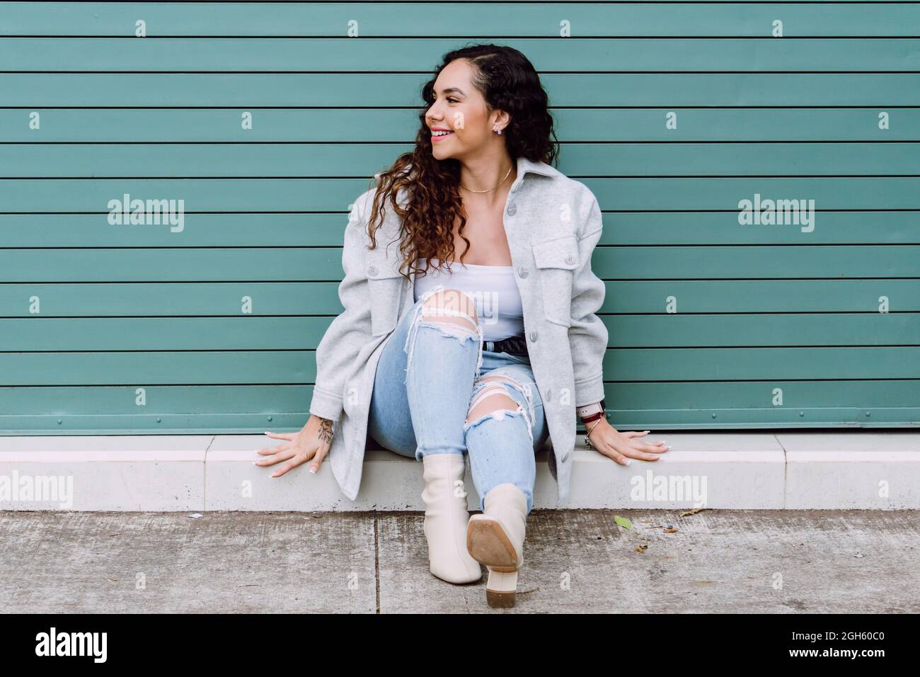 Affascinante donna in abbigliamento elegante seduta vicino alla parete dell'edificio, sorridendo e guardando via Foto Stock