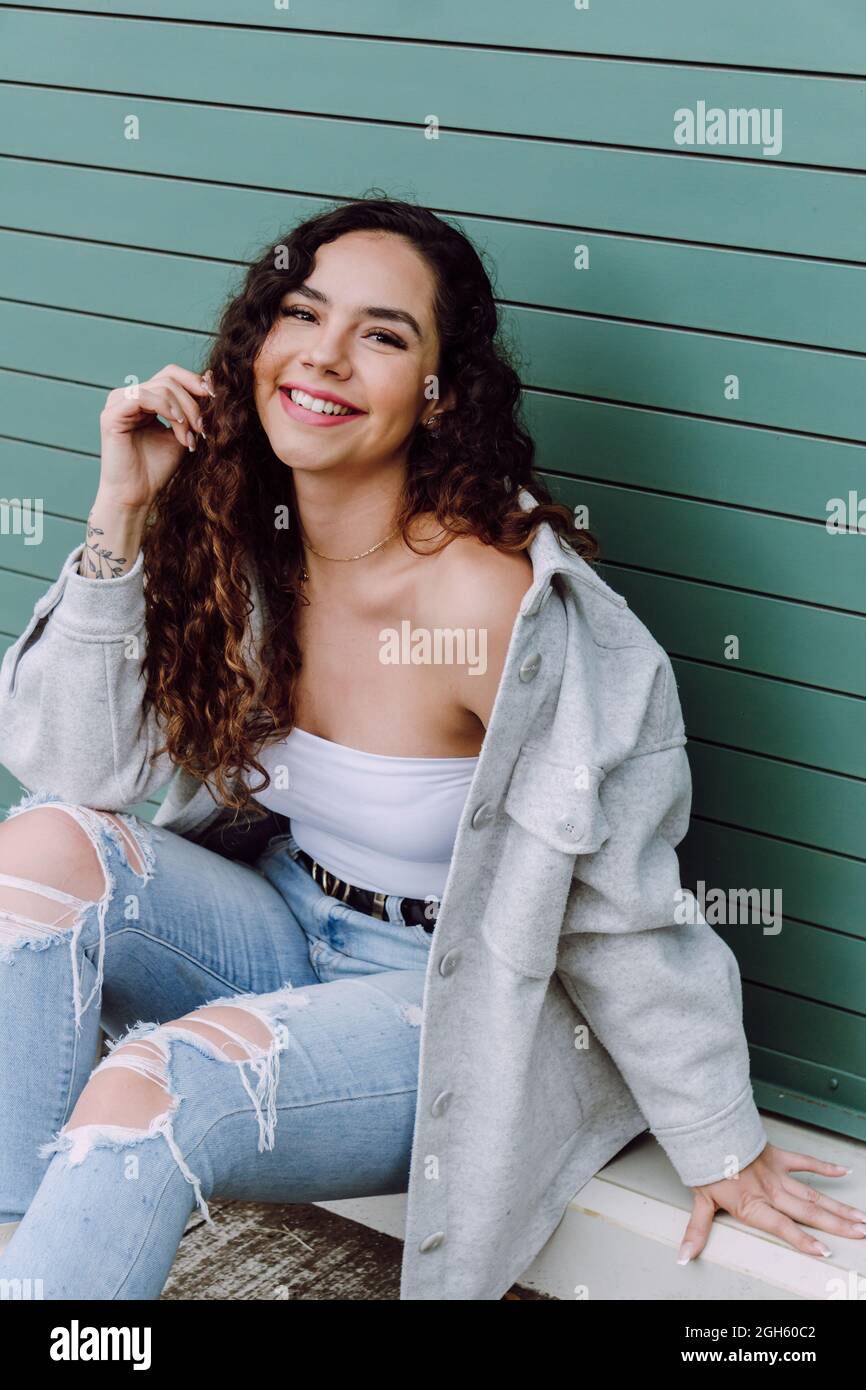 Affascinante donna in abbigliamento elegante seduta vicino alla parete dell'edificio e appoggiata a portata di mano, sorridendo e guardando la macchina fotografica Foto Stock