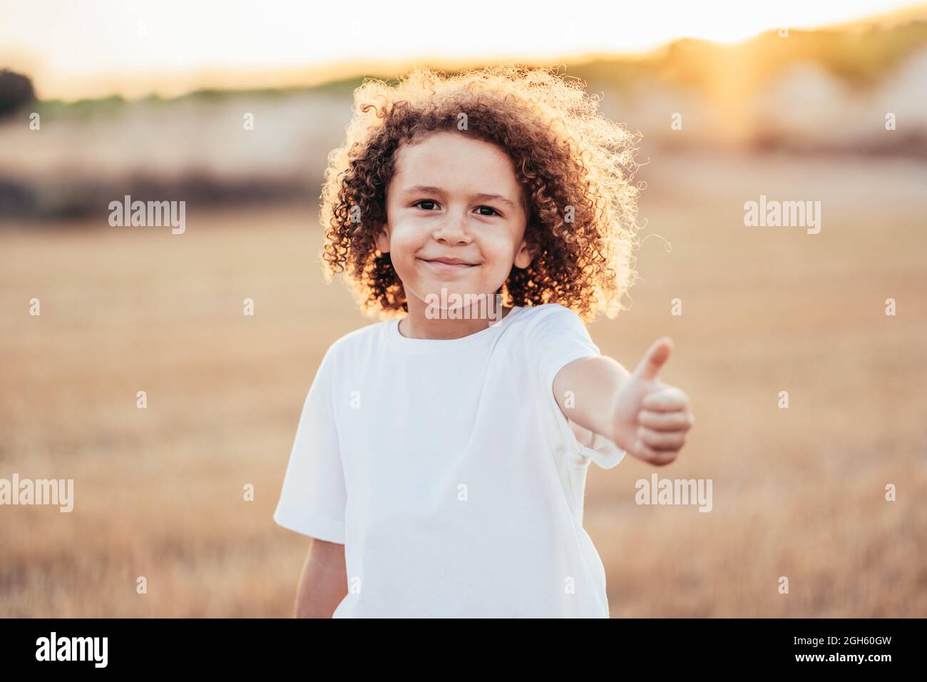 Bambino allegro etnico con capelli ricci che mostrano come un gesto mentre si trova in campo asciutto in estate in retroilluminazione e vondolando alla macchina fotografica Foto Stock