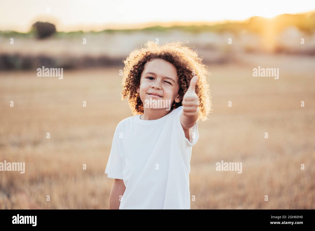 Bambino allegro etnico con capelli ricci che mostrano come un gesto mentre si trova in campo asciutto in estate in retroilluminazione e vondolando alla macchina fotografica Foto Stock