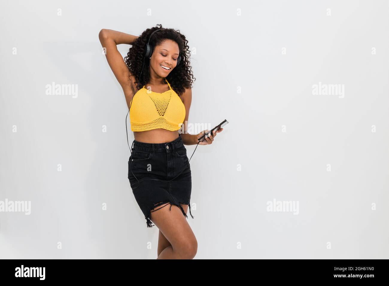Happy African American femmina in cuffie con cellulare danzare mentre si ascolta la canzone su sfondo chiaro Foto Stock