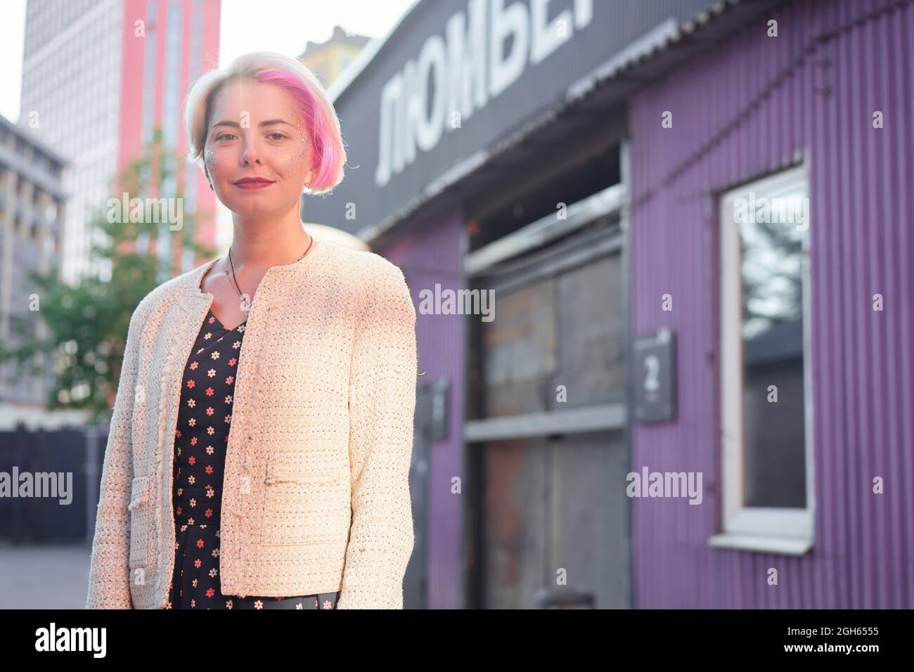 Affascinante alternativa femminile con capelli tinti e in abiti alla moda in piedi contro la strada in città e guardando la macchina fotografica Foto Stock