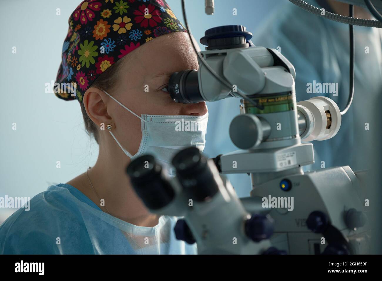 Medico adulto focalizzato in maschera sterile e cappuccio medico ornamentale guardando attraverso microscopio chirurgico contro il coltivatore in ospedale Foto Stock