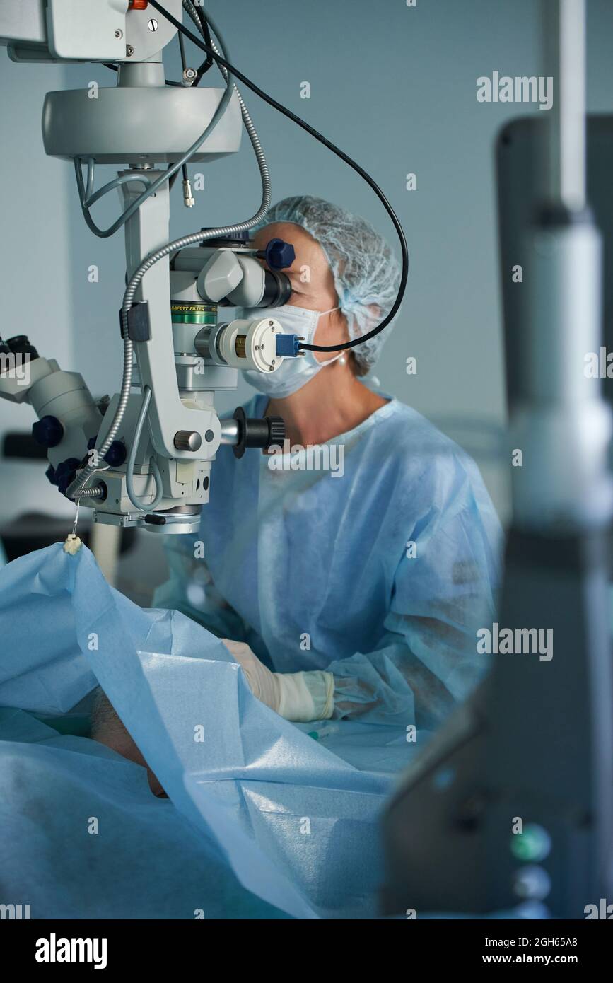 Medico adulto focalizzato in maschera sterile e cappuccio medico ornamentale guardando attraverso microscopio chirurgico contro il coltivatore in ospedale Foto Stock