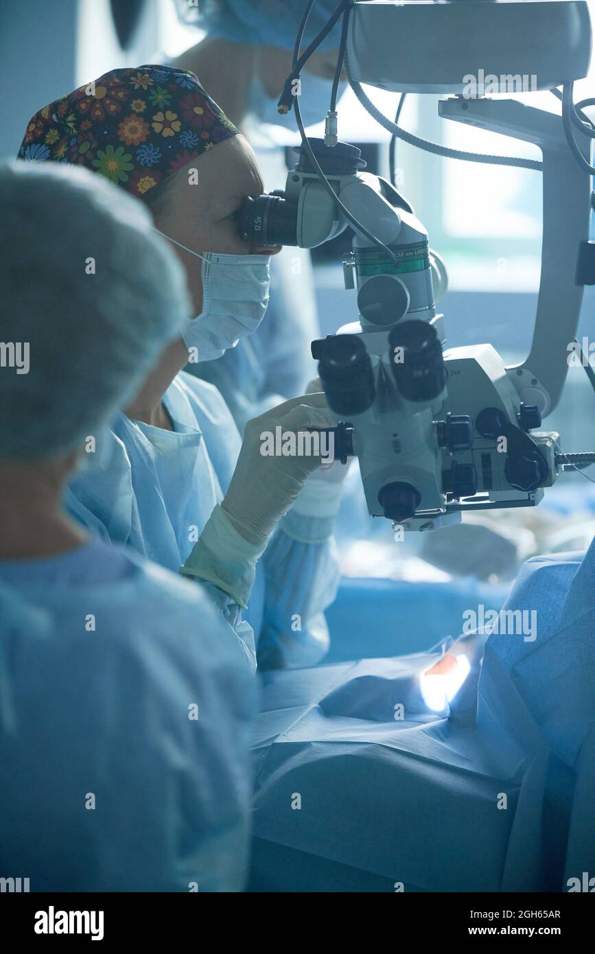 Medico femminile adulto in maschera sterile e cappuccio medico ornamentale guardando attraverso il microscopio chirurgico contro il coltivatore in ospedale Foto Stock