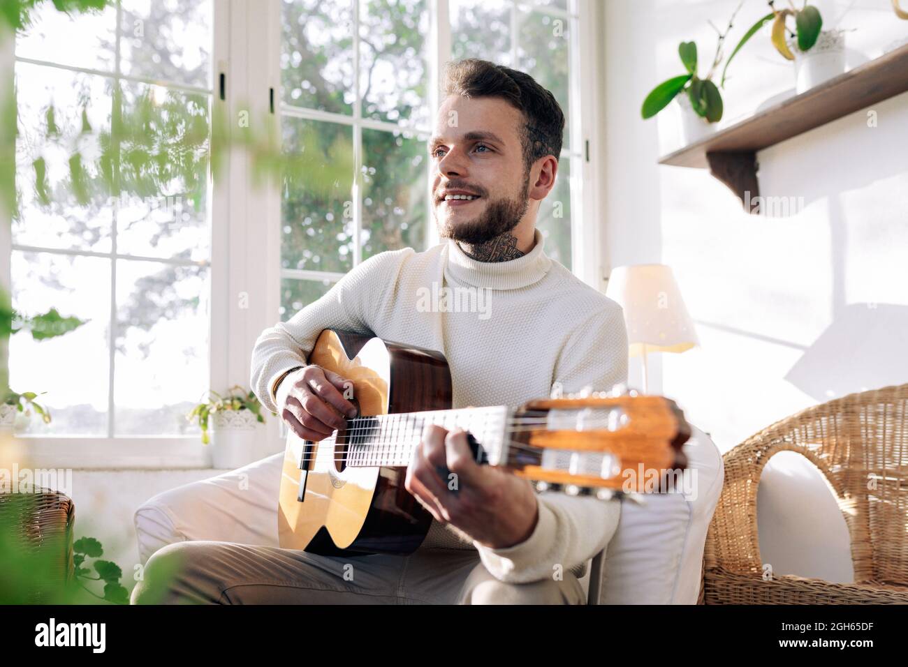 Musicista maschile contemplativo con tatuaggi che suonano chitarra classica  mentre si siede in poltrona e si guarda lontano contro la finestra in casa  Foto stock - Alamy