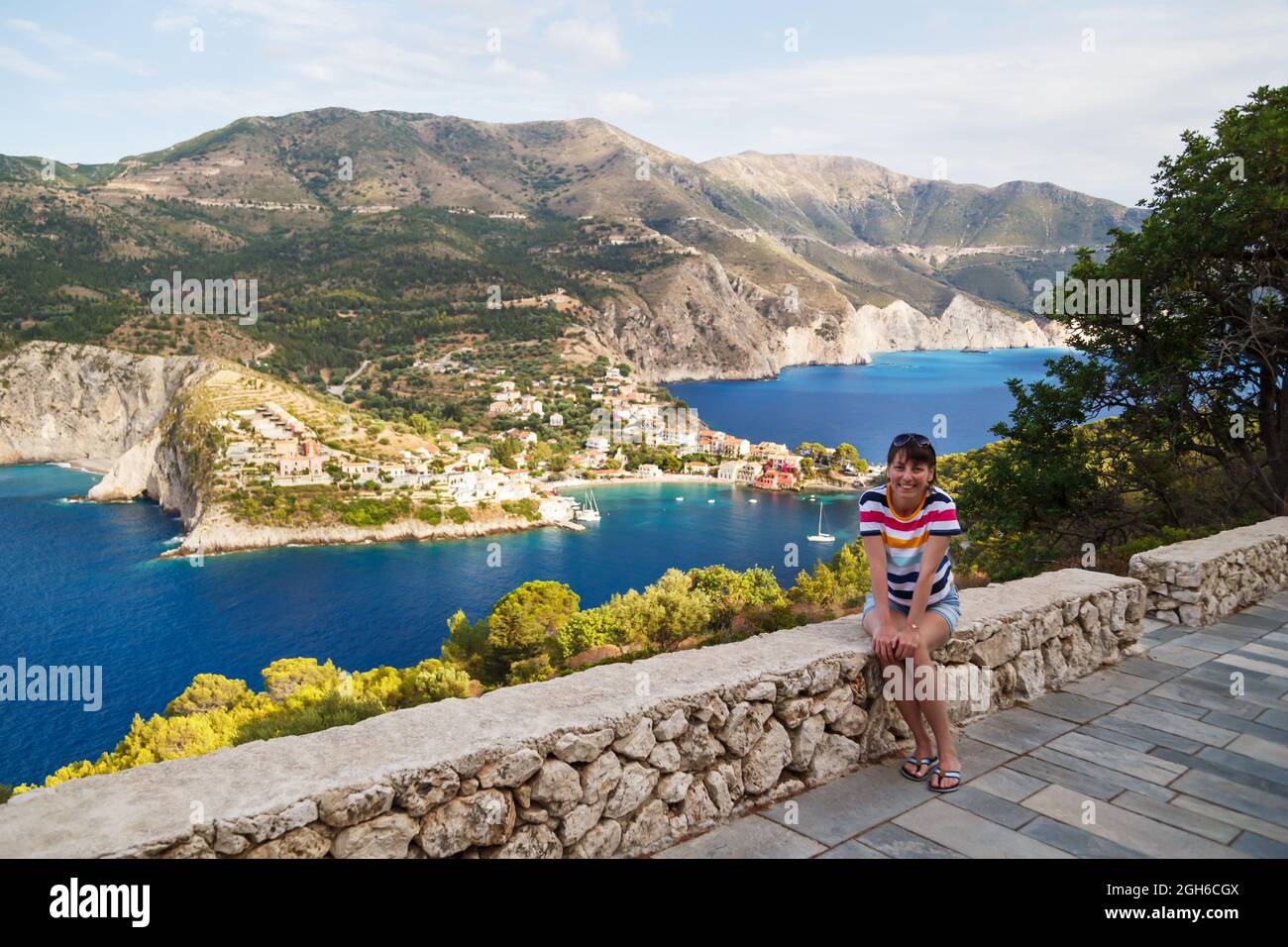 Giovane donna sorridente turista in maglia multicolore a righe sulla cima della montagna sulla baia blu di Asos. Vista aerea, scenario estivo di destinazione turistica estremamente popolare a Cefalonia, Grecia, Europa Foto Stock