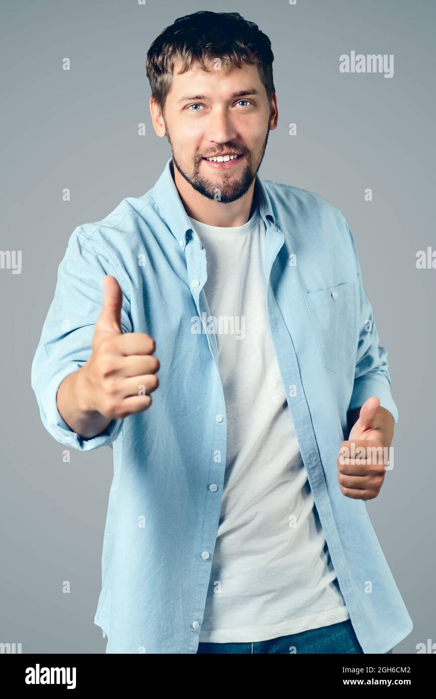 Ritratto di un felice uomo caucasico sorride e mostra i suoi pollici in su. Un ragazzo positivo in abiti casual mostra un simile su uno sfondo grigio. Foto Stock