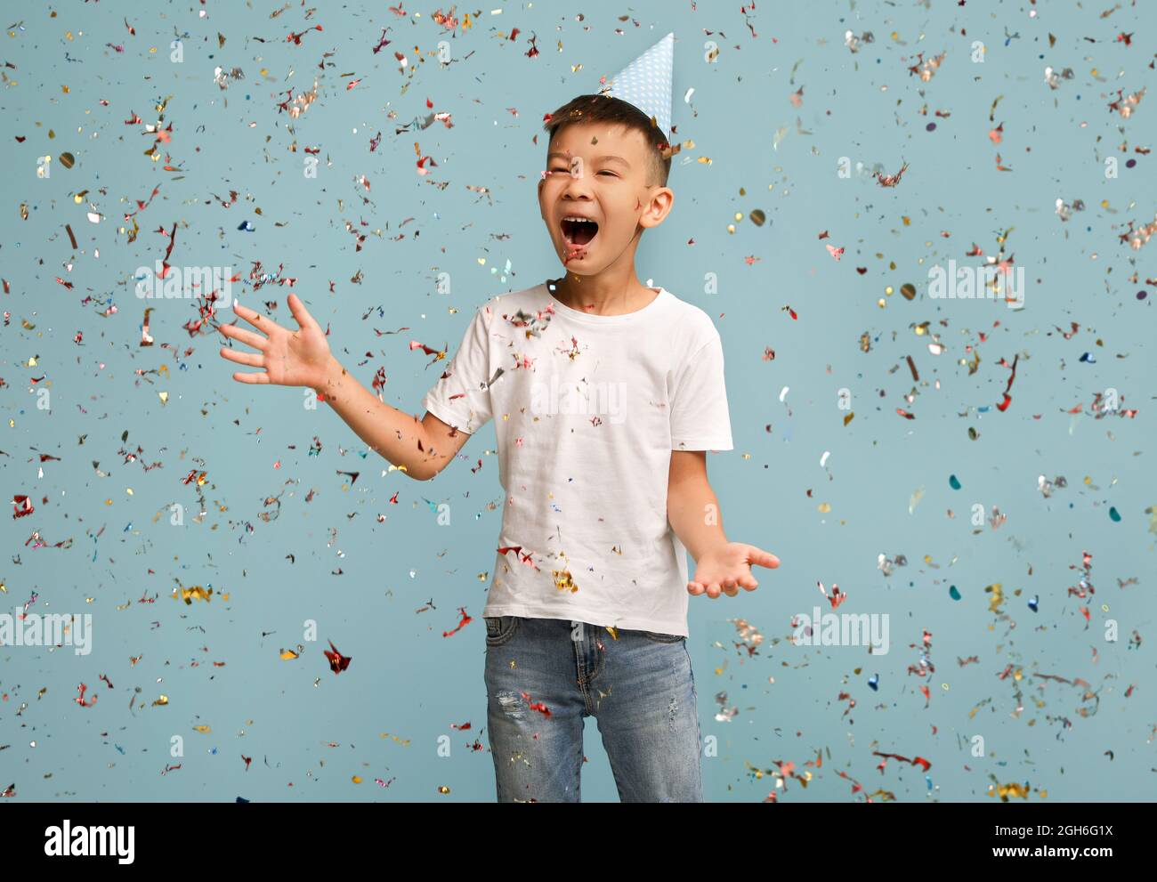 Festa di compleanno. Buon ragazzo asiatico che indossa cappello B-Day in piedi sotto Falling Confetti Foto Stock