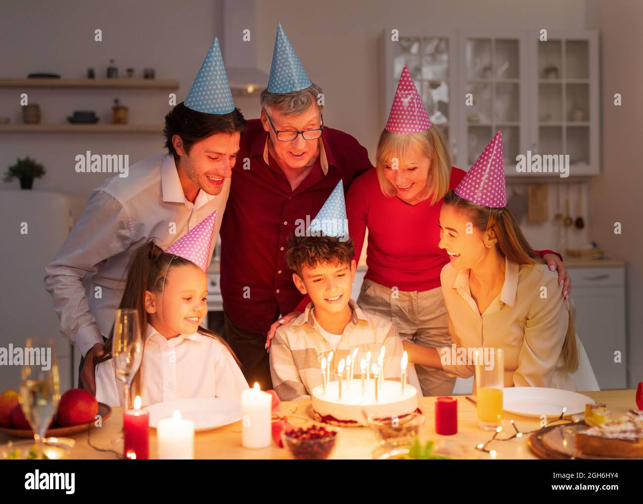 Ragazzo allegro che riceve torta di compleanno con candele brucianti dalla famiglia premurosa, celebrando le vacanze al chiuso di notte Foto Stock