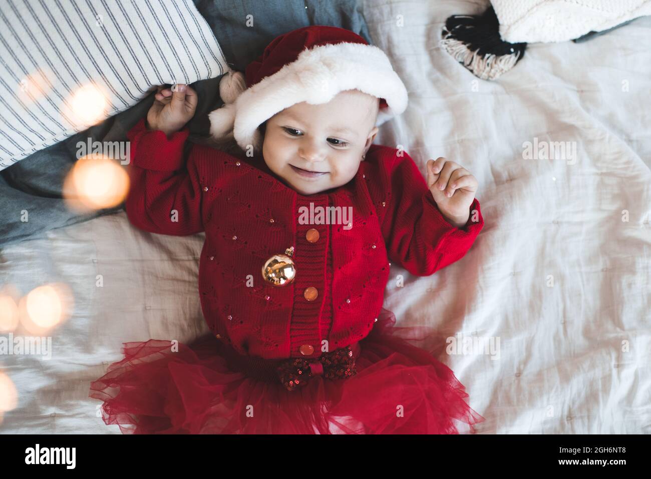 Cute baby girl 1 anno vecchio indossare cappello babbo natale rosso e abito a maglia sdraiato nel letto da vicino. Bambino sorridente che festeggia le vacanze di natale. Vista dall'alto Foto Stock