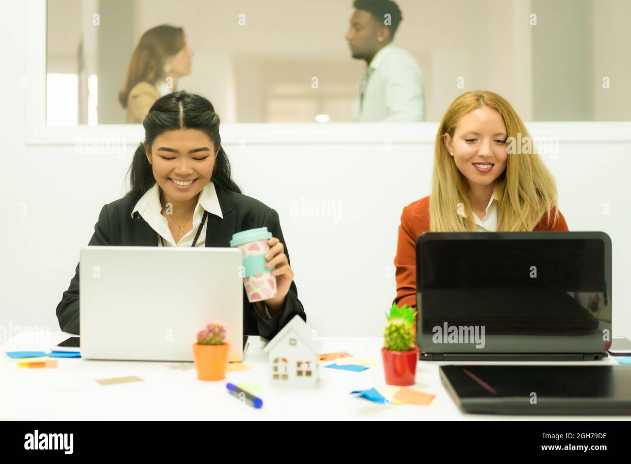 Foto di allegre donne multirazziali in camicia bianca e giacca nera, sorridenti e scriventi con un computer portatile. Concetto di lavoro e studio. Foto Stock