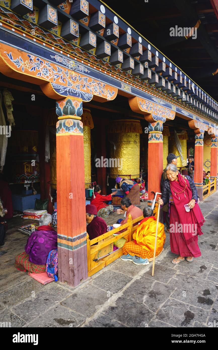 Una donna bhutanese anziana in abito tradizionale cammina accanto a colonne dipinte con colori vivaci sui terreni del National Memorial Chorten a Thimphu, Bhutan Foto Stock