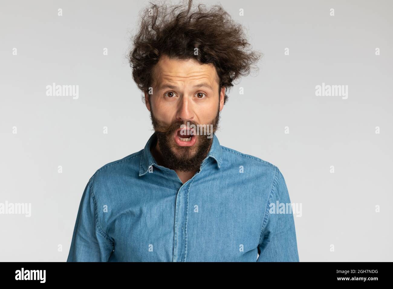 giovane uomo temuto in denim camicia con la barba lunga apertura bocca e gridando mentre si posa su sfondo grigio in studio Foto Stock