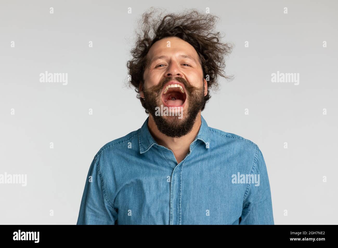 giovane uomo felice in denim camicia con barba lunga divertirsi e ridere, urlare e posare su sfondo grigio in studio Foto Stock