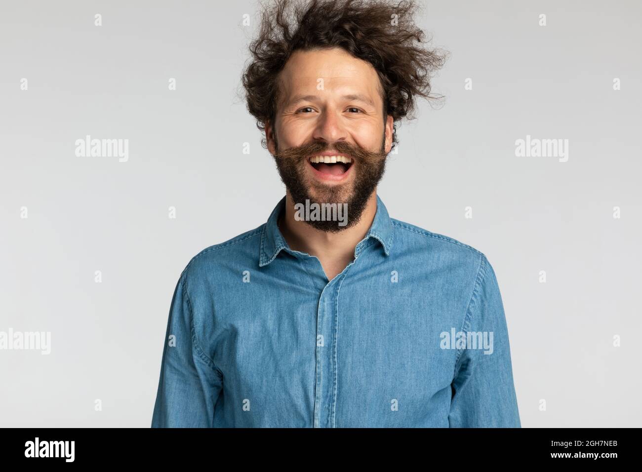 ritratto di felice riccio ragazzo capelli in denim camicia con barba lunga e baffi divertirsi e ridere su sfondo grigio in studio Foto Stock