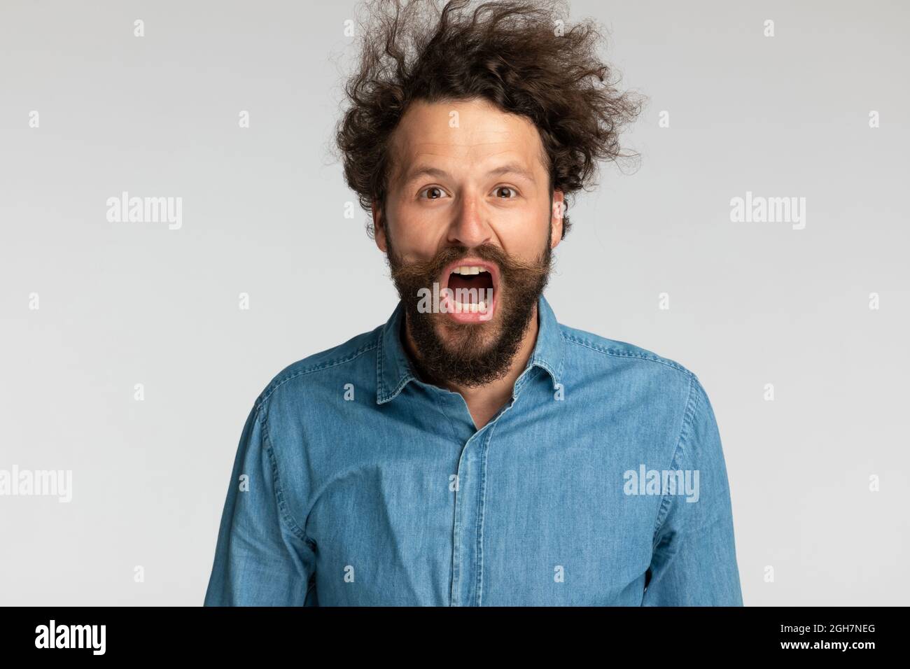 ritratto di giovane uomo sconvolto in denim camicia apertura bocca e gridando su sfondo grigio in studio Foto Stock