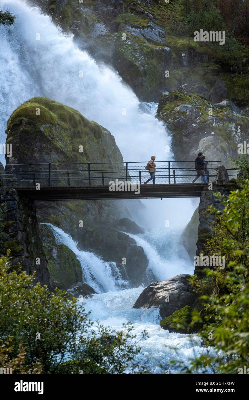 I turisti attraversano un ponte di fronte a una cascata infestante al ghiacciaio Briskdal, al parco nazionale del ghiacciaio Jostedal, Norvegia, Europa Foto Stock