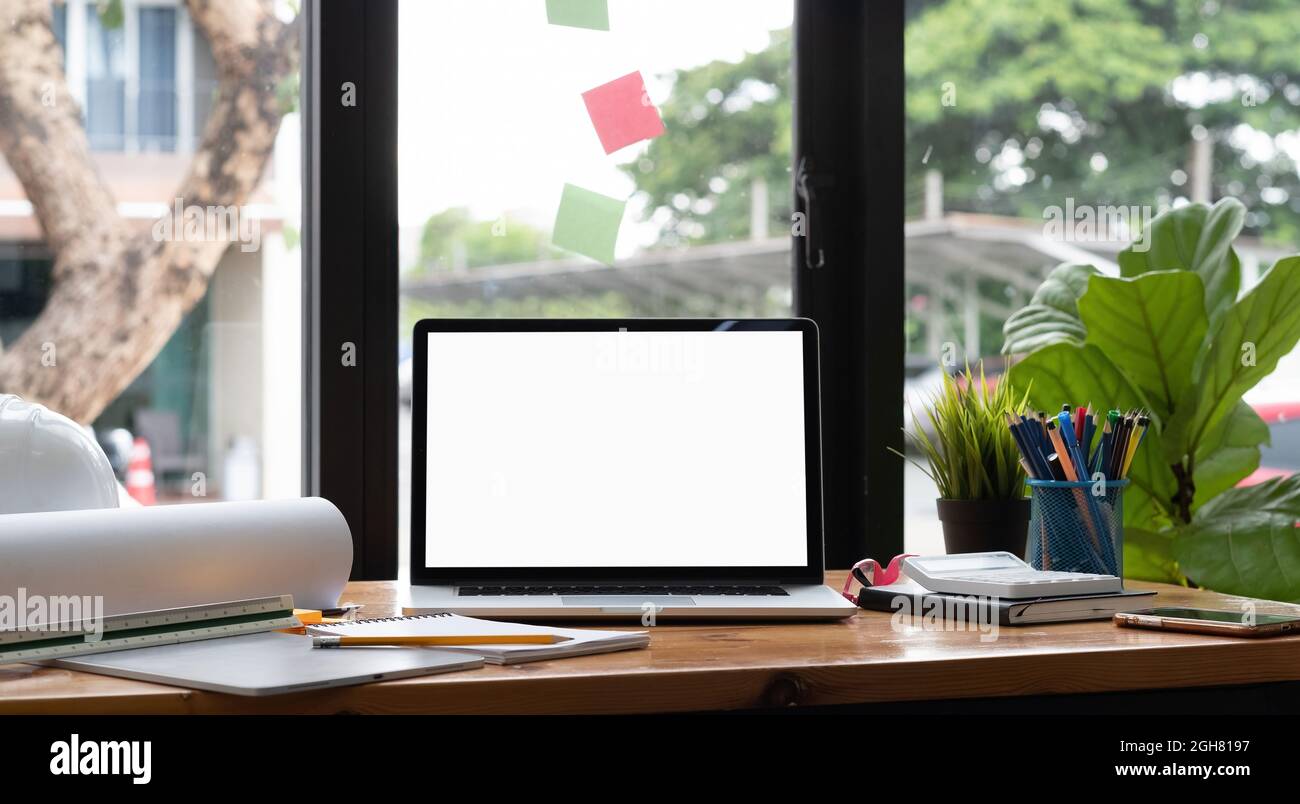 Computer portatile con schermo bianco su scrivania in legno, spazio di lavoro ingegneristico con disegno di casco di sicurezza. Foto Stock