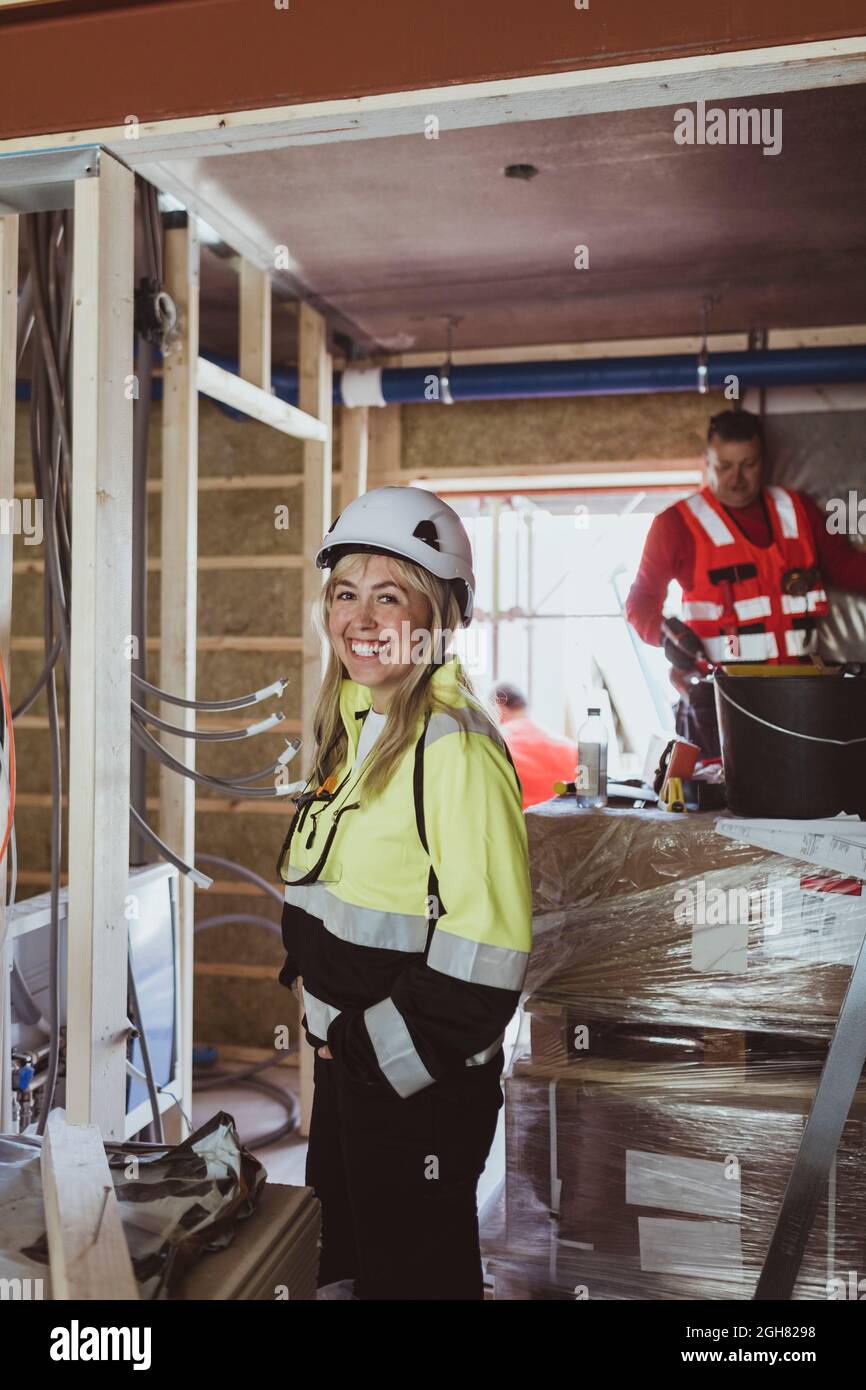 Felice imprenditore di costruzione femminile in piedi con la mano in tasca, mentre il lavoratore di costruzione maschile che lavora in loco Foto Stock