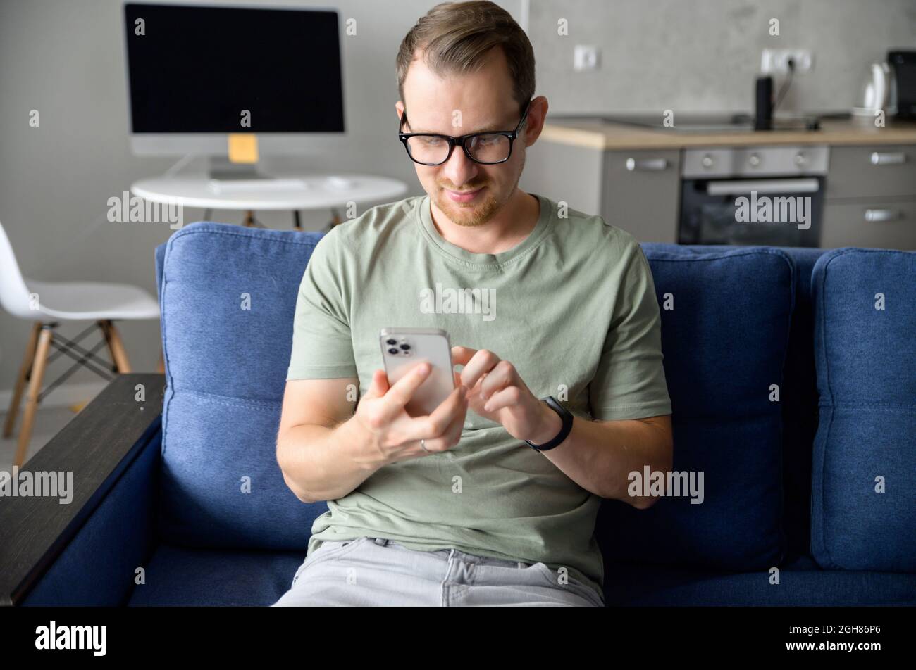 Bel giovane uomo sorridente in occhiali e t-shirt casual testing su smartphone, scorrimento news feed, passa il tempo in social network seduti sul divano in appartamento moderno Foto Stock