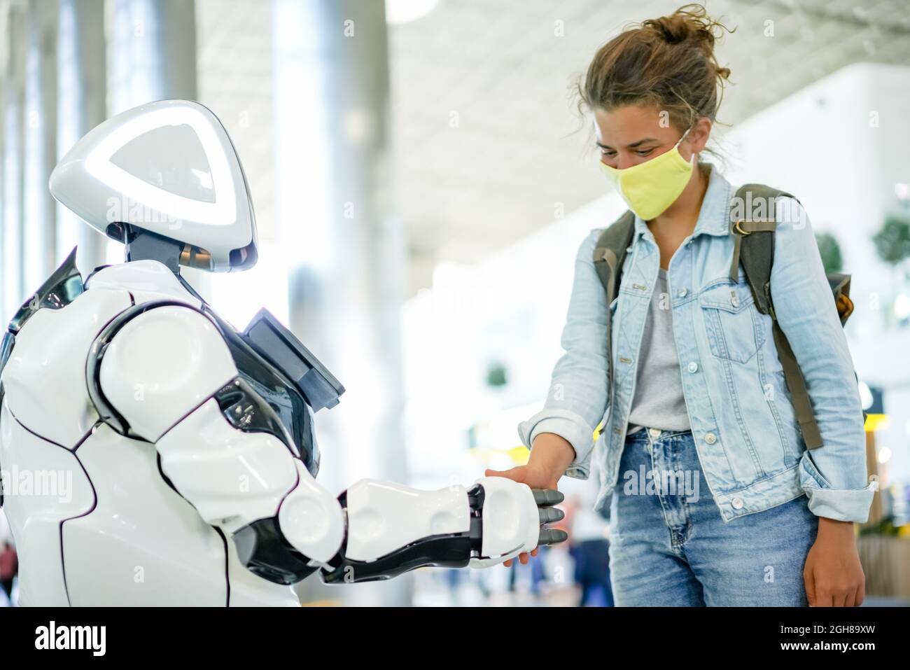 Robot e handshake umano in un luogo pubblico, concetto tecnologico moderno futuro. Assistente robotico intelligente con intelligenza artificiale e graziosa donna in maschera gialla Foto Stock