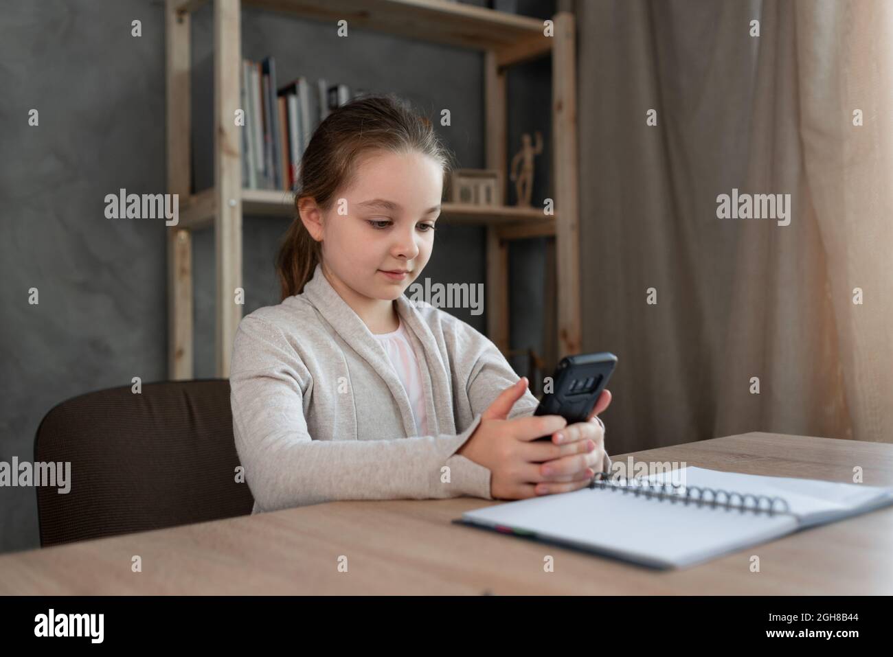 Prenditi una pausa dall'esplorazione di una bambina caucasica seduta a un tavolo nel corridoio utilizzando il suo smartphone e divertendosi, Foto Stock