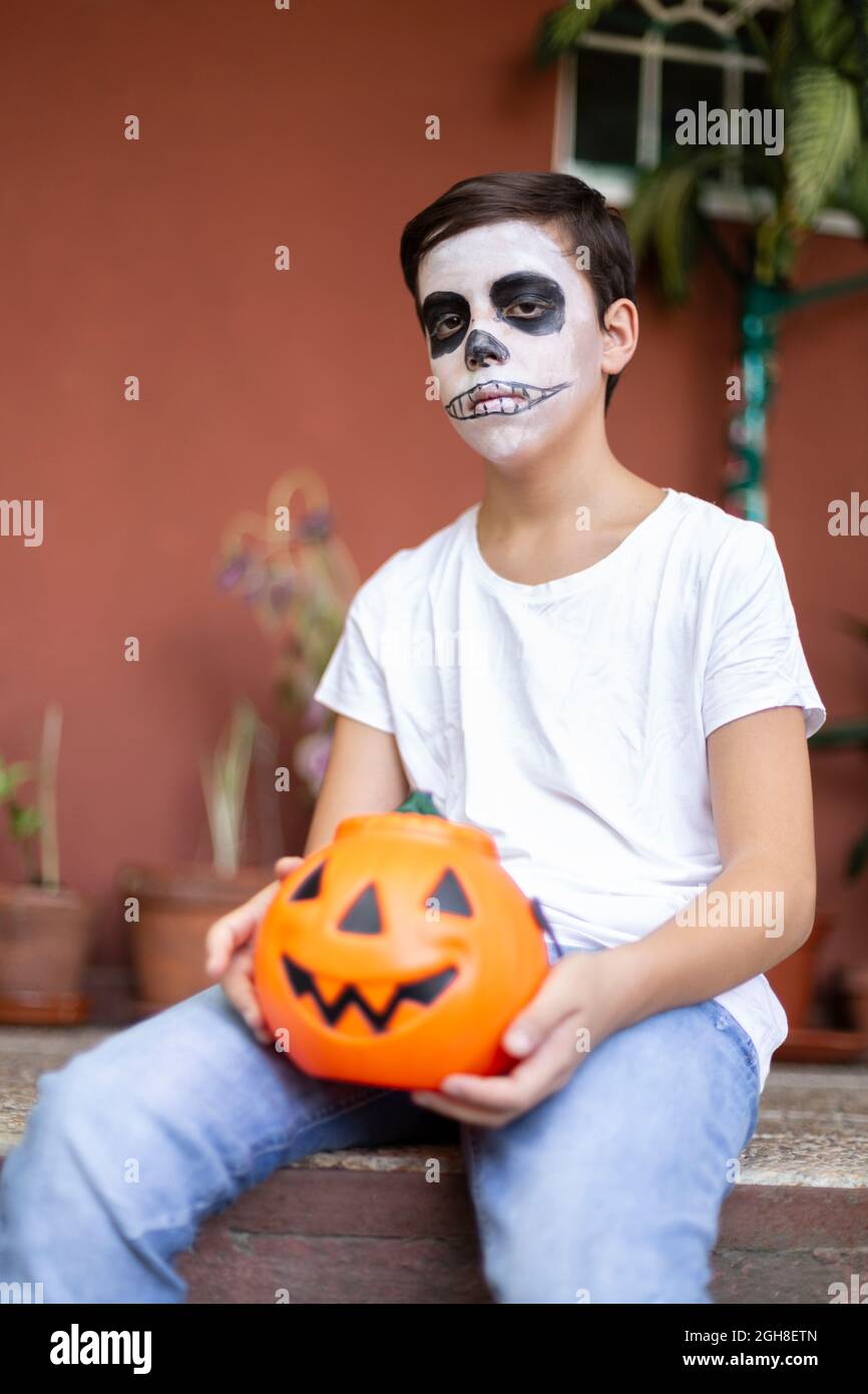 Ritratto di ragazzo con il viso fatto come un teschio seduto all'ingresso della sua casa. È molto serio e tiene una zucca di Halloween. Foto Stock