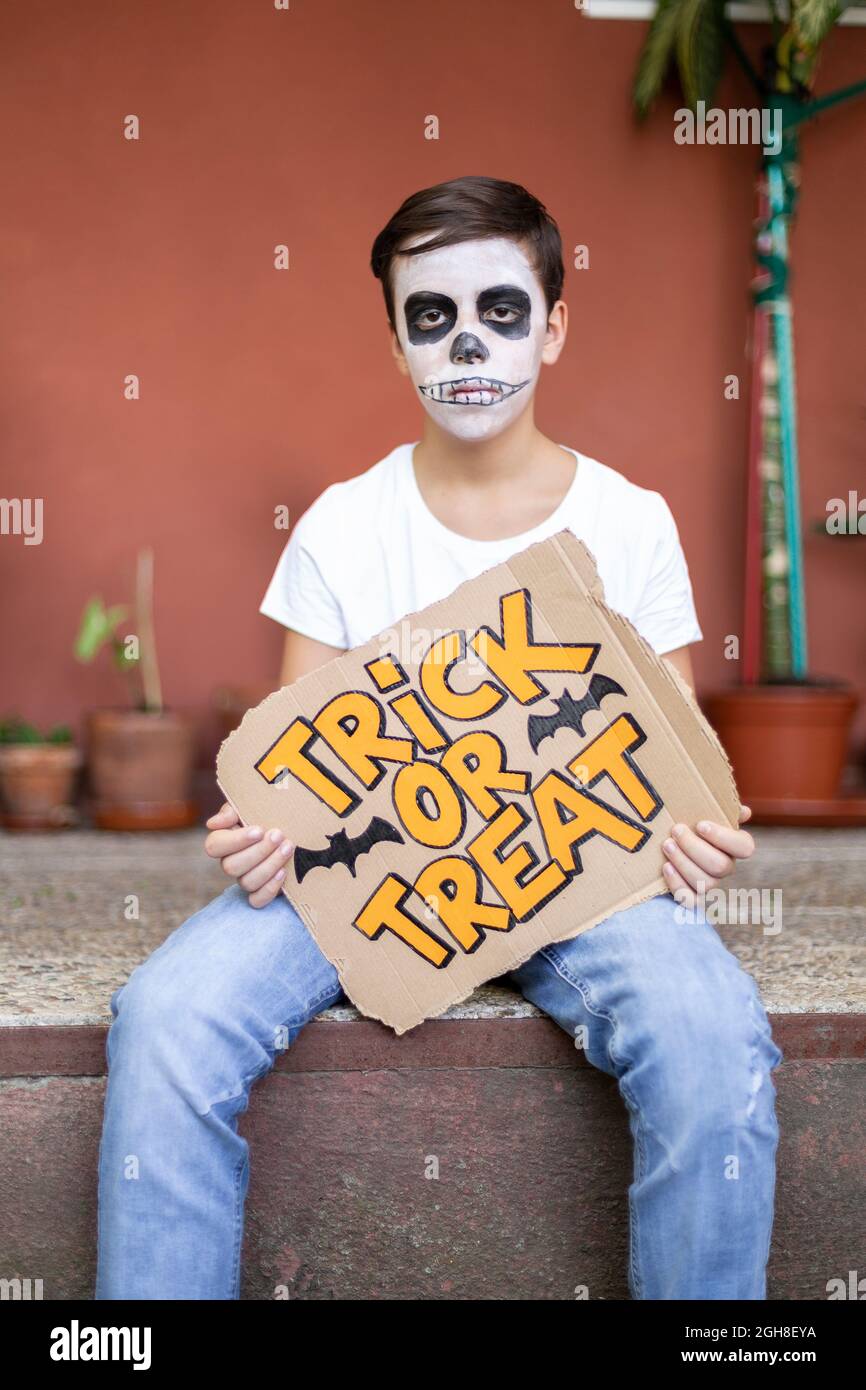 Ragazzo adolescente con volto caratterizzato da un cranio seduto all'aperto. È serio e tiene nelle sue mani un segno con il testo Trick or Treat. Halloween. Foto Stock