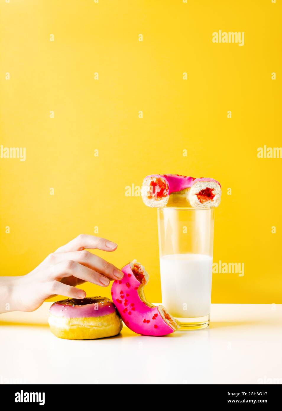 Mano bianca donna che raggiunge per una dolce ciambella rosa con marmellata di bacche e bicchiere di latte vegetale. Caffetteria o caffetteria con pasta di zucchero fresco, ricco di calorie Foto Stock