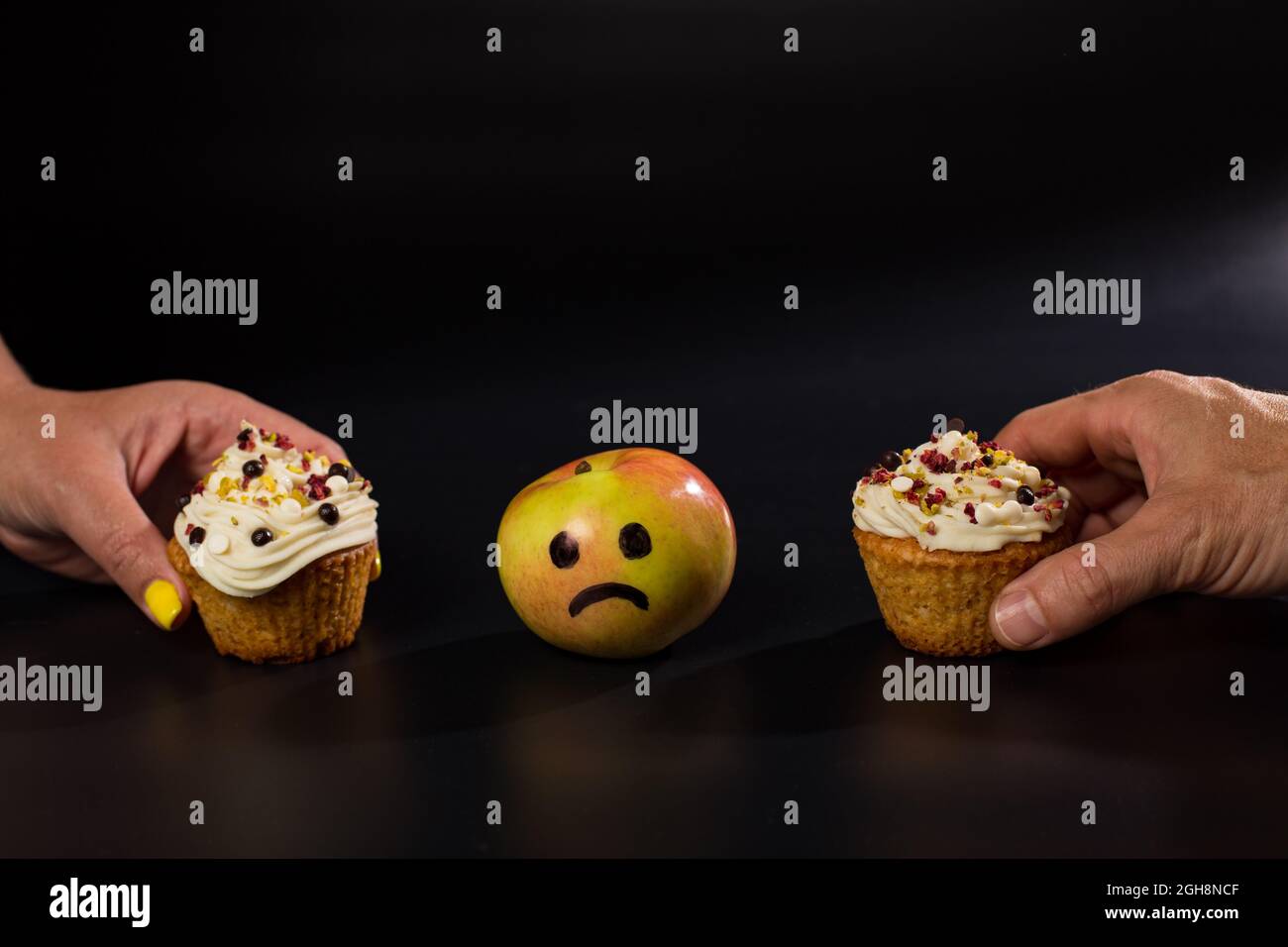 Due mani che soffocano i cupcake dolci contro la mela fresca, su sfondo nero. Foto Stock