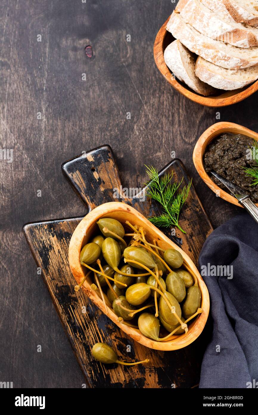 Capperi in scatola in piatto di legno, tapenade e pane su un vecchio sfondo alla moda di legno scuro. Vista dall'alto Foto Stock