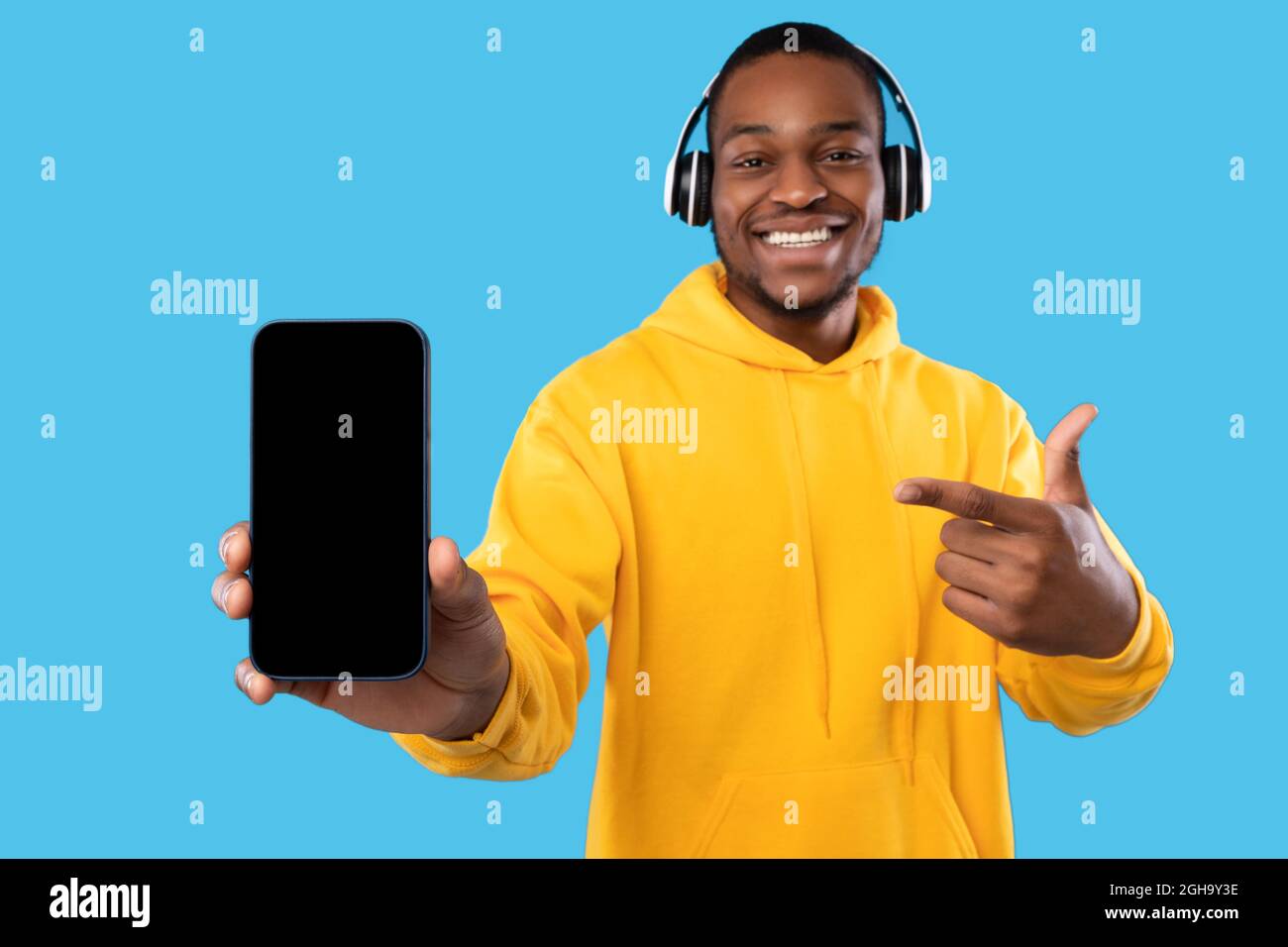 Uomo nero che mostra il telefono schermo vuoto indossando cuffie, sfondo blu Foto Stock