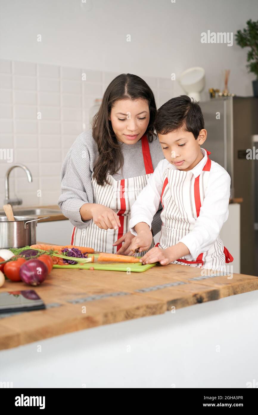 Il bambino impara a tagliare le verdure mentre la madre lo guarda e lo aiuta con attenzione. La gente latina che fa il cibo tipico latino-americano. Madre e figlio Foto Stock