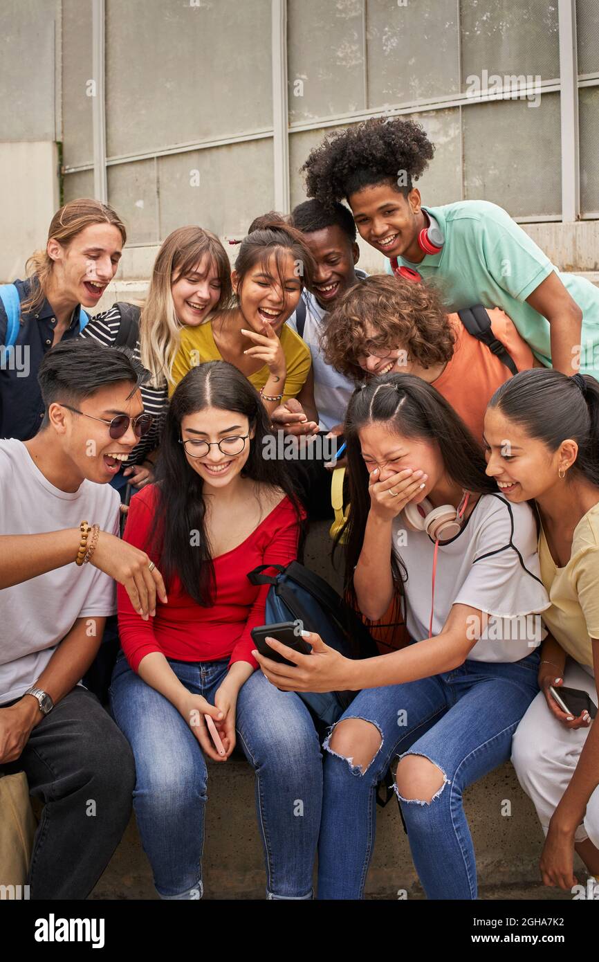 Foto verticale di un gruppo multietnico di studenti che usano il cellulare e ridono. Gli adolescenti usano uno smartphone, navigano sul Web. Divertirsi insieme. Foto Stock