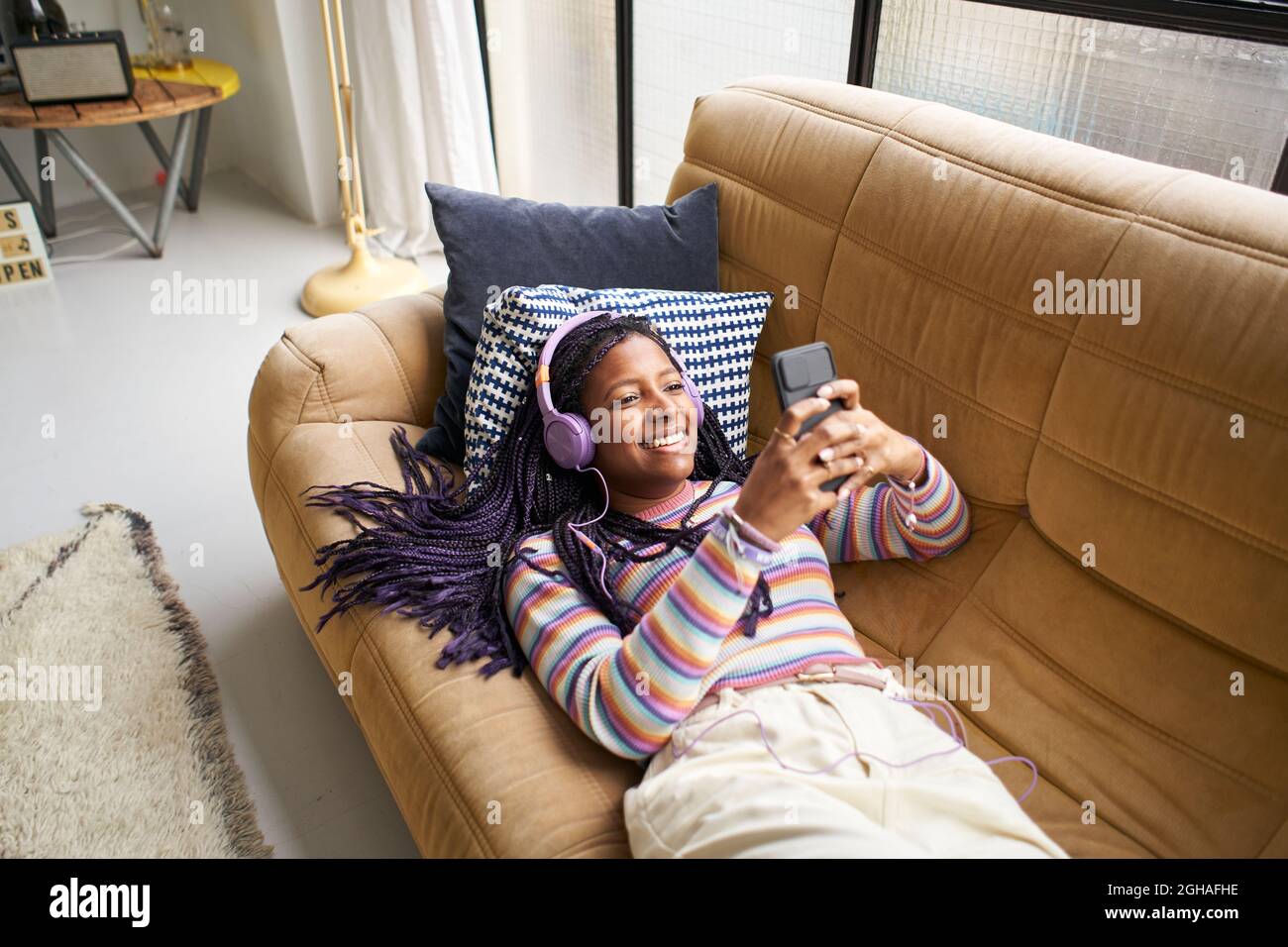 Sorridente donna americana che ascolta la musica con le cuffie e utilizzando lo smartphone rilassarsi sul divano a casa.concetto di gioventù e tempo libero. Foto Stock